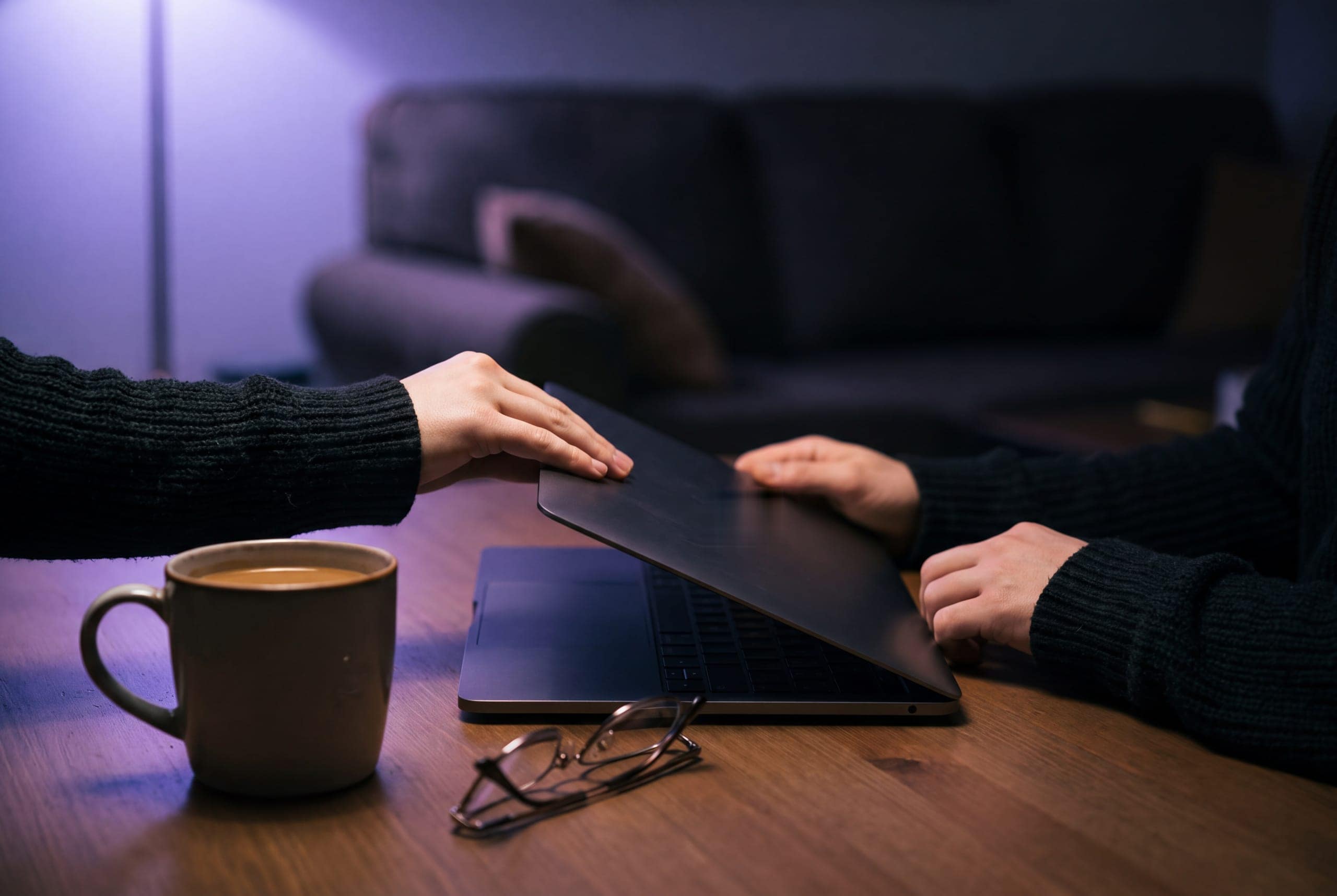 A laptop being closed on a table beside a mug, lit by a lamp