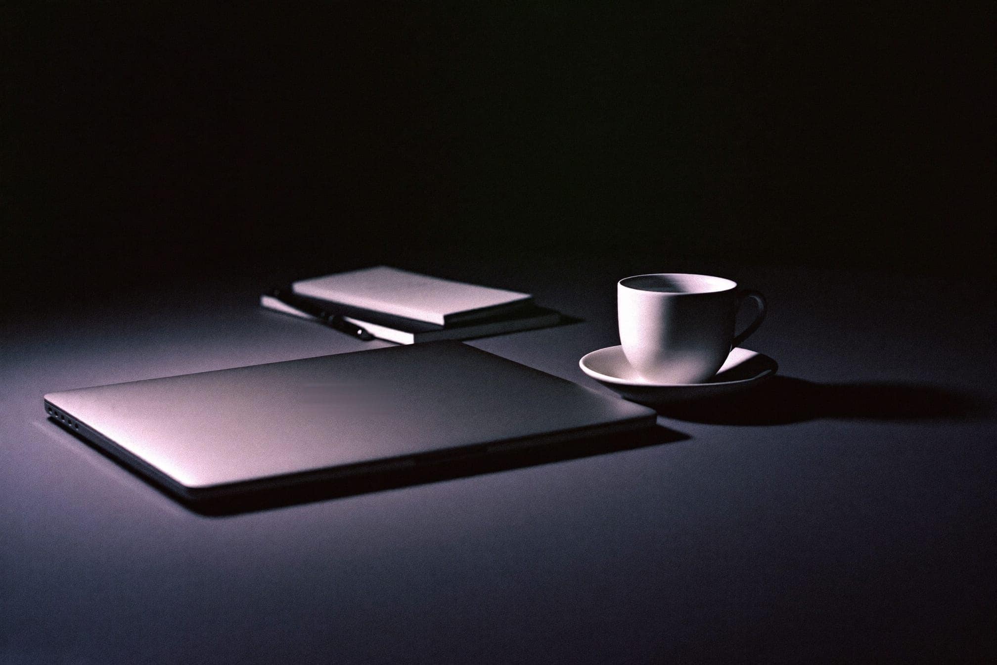 A closed laptop, a notebook and a cup on a desk in low light
